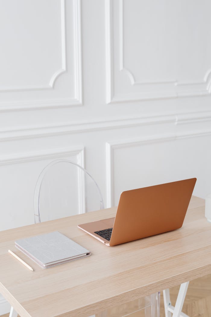 A modern minimalist workspace featuring a sleek laptop and notebook on a wooden desk against a white paneled wall.